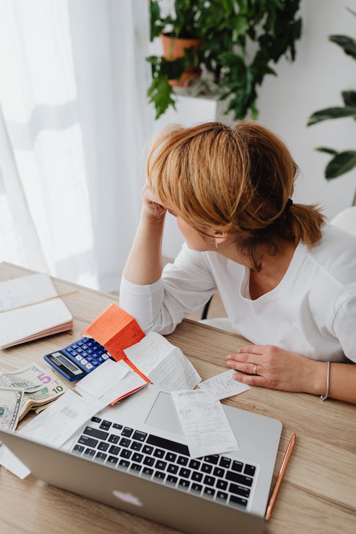 A woman overwhelmed by bills and finances sits at a desk with a laptop and calculator.